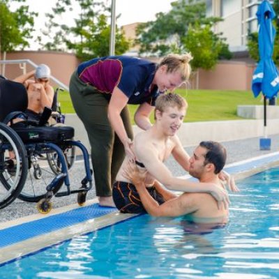 A young man is being helped from a wheelchair into a swimming pool by two trainers.